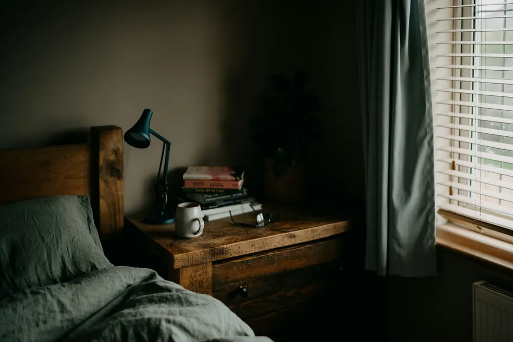 A dim bedroom with green linen bedding, a wooden nightstand with a lamp and books, and a curtain beside the window