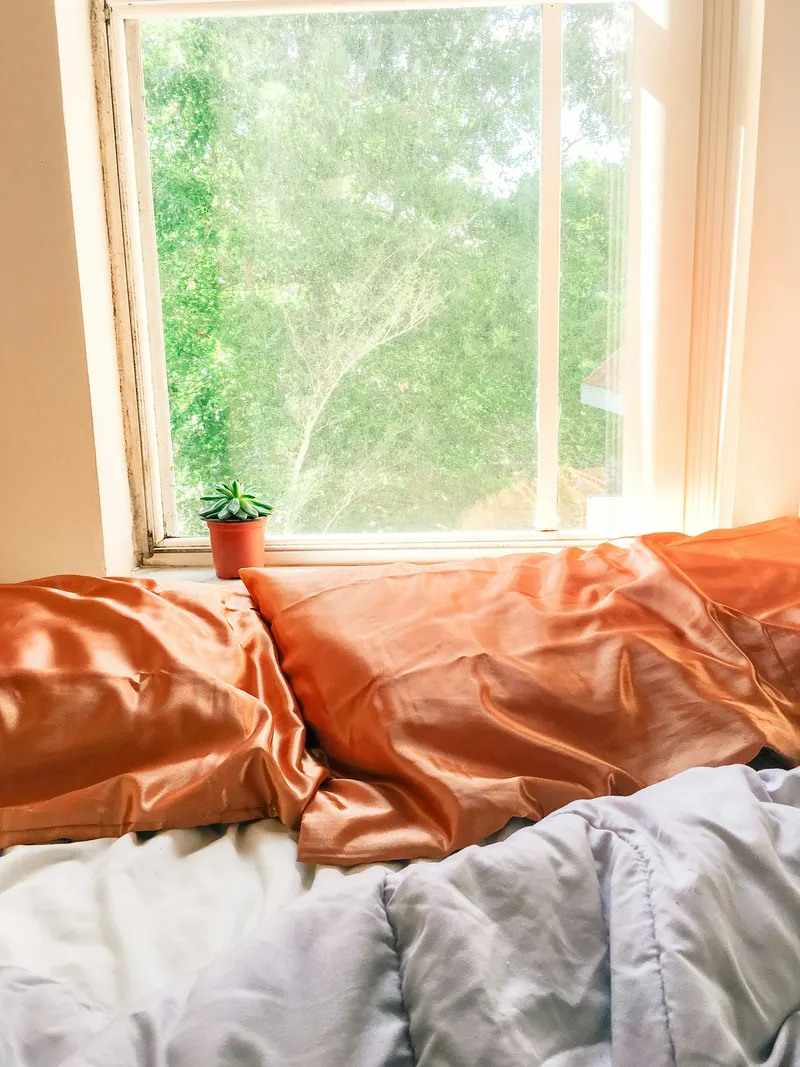A bed with orange satin pillowcases and a lavender comforter beside a sunny window with green trees outside