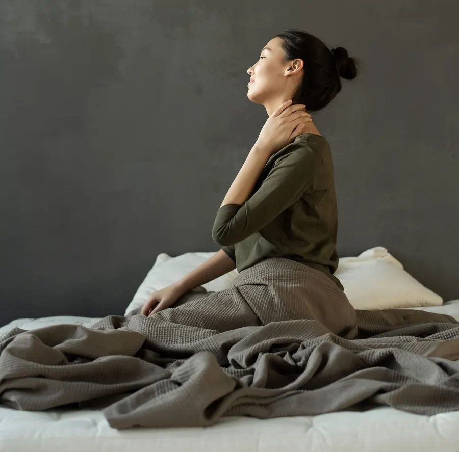 A woman sitting up in bed in the morning with one hand on the back of her neck