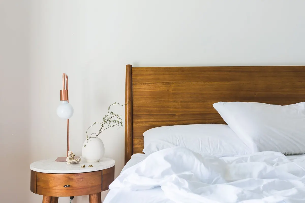 A wooden bed with white bedding and pillows beside a round nightstand with a lamp, a bed that needs a 3-foot clear zone around any heater