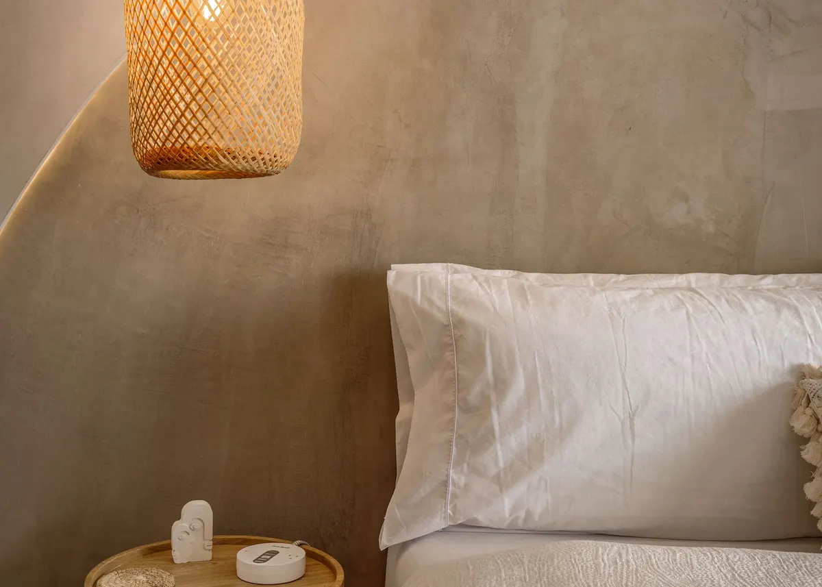 A calm bedroom with a woven pendant lamp, a round wooden side table and a bed with white pillows