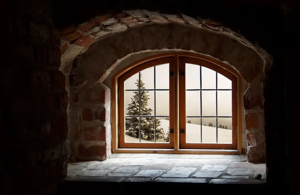 An old arched wooden window set in a thick stone wall, looking out on a snowy hillside with a pine tree