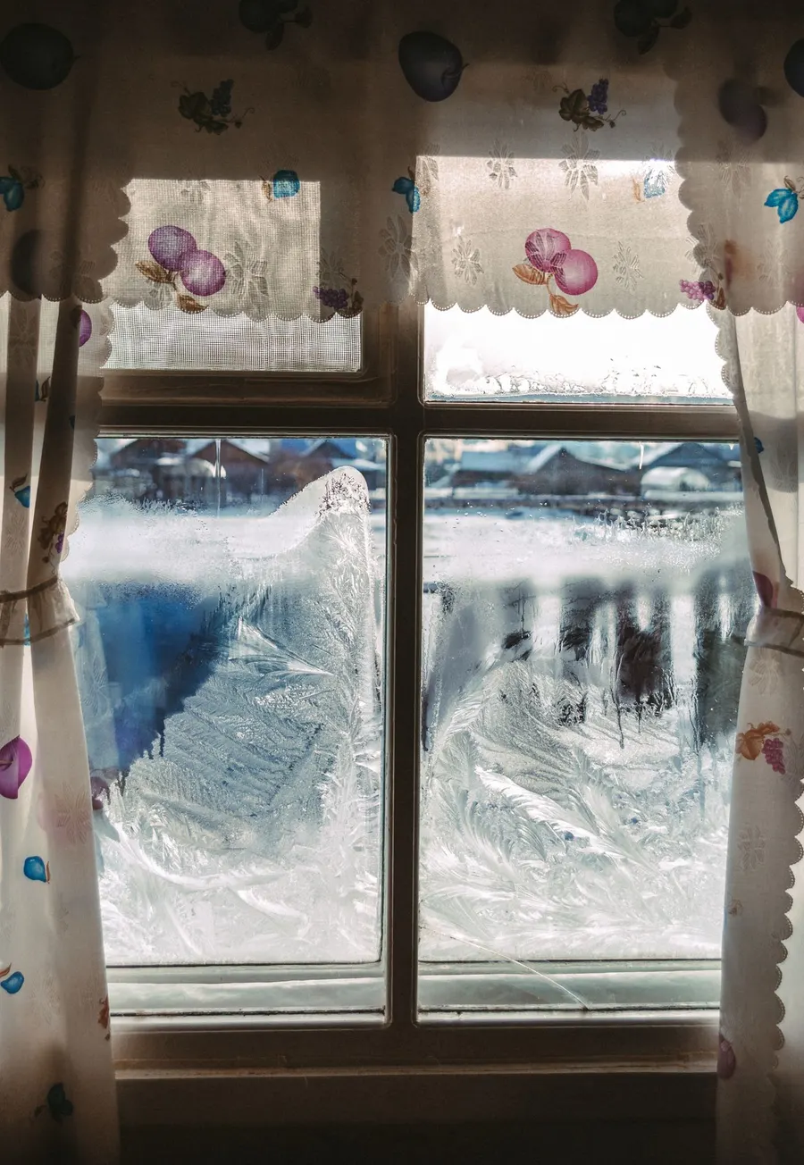A frosted window with floral curtains on a freezing winter morning