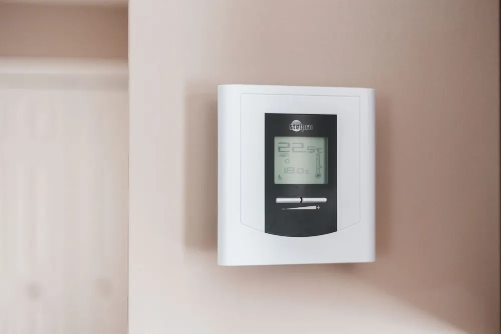 A white digital programmable thermostat mounted on a light pink wall