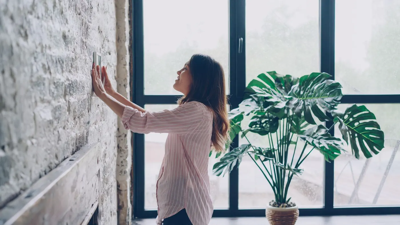 Hand adjusting a wall thermostat in a bright living room — the same instinct that decides between a cooler and an AC
